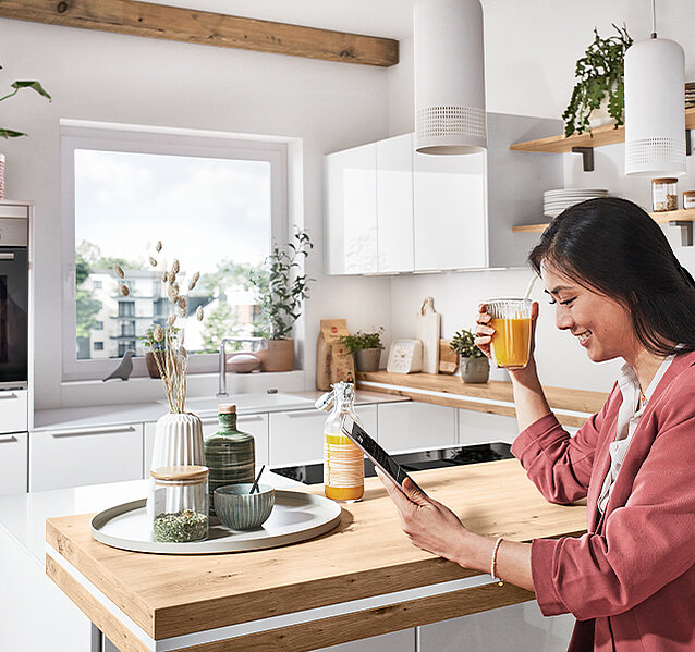 Eine Frau genießt ihren Morgen Tee, während sie auf einem Tablet in einer modernen, hellen Küche mit stilvollen Holzakzenten und grünen Pflanzen surft.